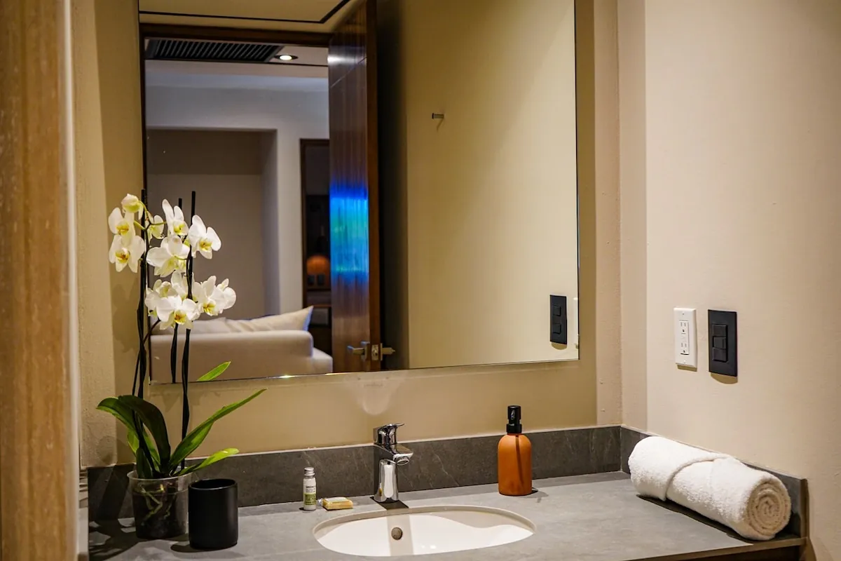 Baño del departamento con lavabo de piedra, espejo iluminado y una orquídea blanca